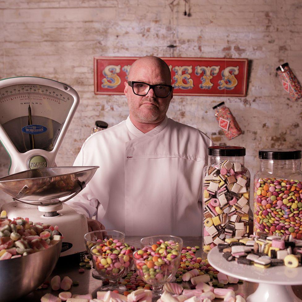 Heston Blumenthal wearing a white chef's outfit, surrounded by sweets