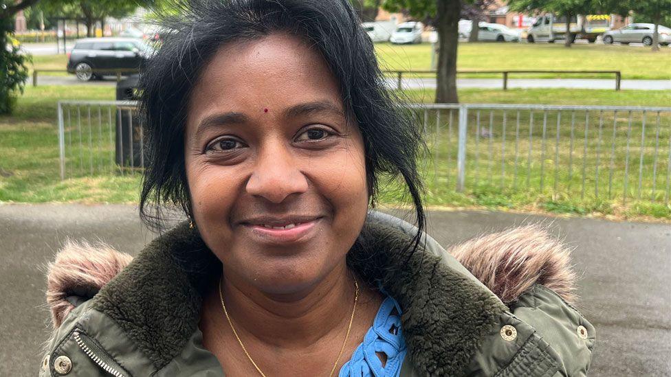 Anne Benedict who has black hair pulled back from her face and is smiling. She has a light green coat on with a furry collar over a blue top. Behind her is pavement and a railing in front of a park.