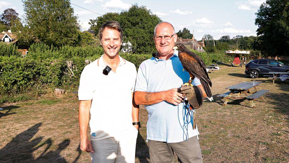 Chesney Hawkes, wearing a white polo shirt, stands next to Wayne Housden, who is wearing a pale blue polo shirt and a falconer's glove. Both are smiling at the camera. A Harris's hawk is perched on the glove. 
