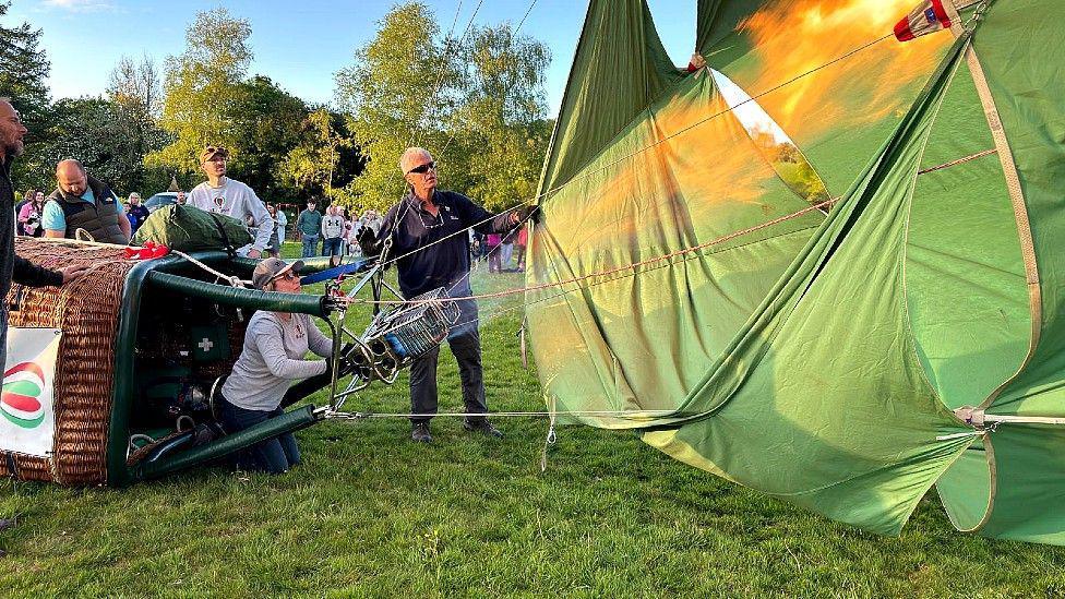A wicker balloon basket is on its side attached to the bottom of green balloon fabric. Laura Davies in on her knees on the ground between the supports of the basket which attach to the balloon, directing a gas heater into the interior of the balloon envelope. She wears jeans, a grey long-sleeve top and a baseball style cap and sunglasses. Three men stand to the far side of her and the basket. A crowd of people can be seen in the background watching her inflate the balloon.