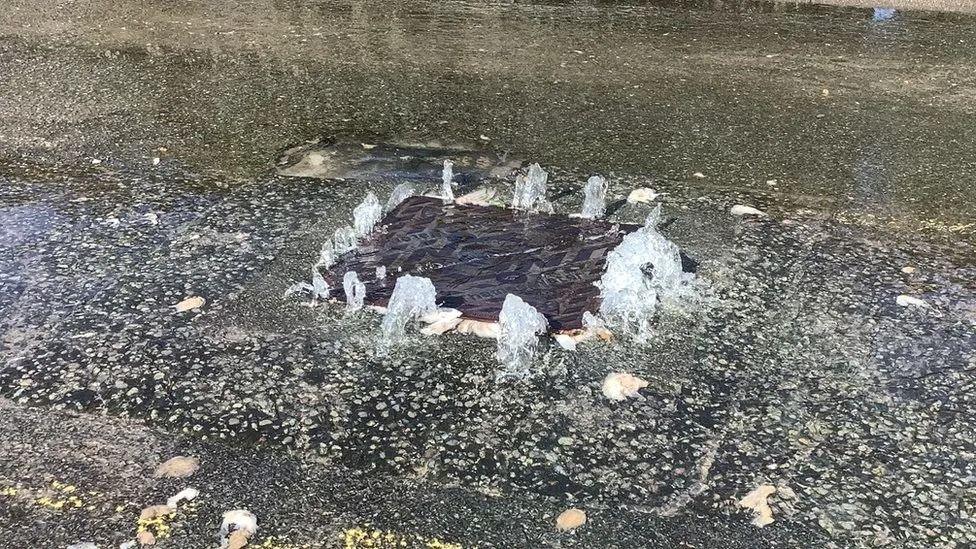 A manhole cover in the middle of a road with water coming up around the edges and flooding the road, with bits of toilet paper floating in it