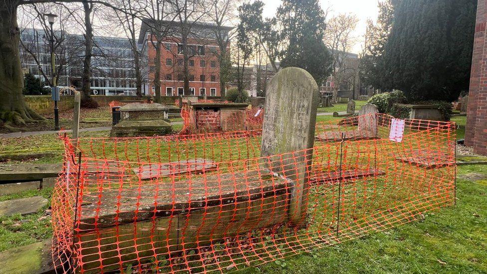 Orange net-style fencing around several stone tombs in churchyard with large office-style building in background