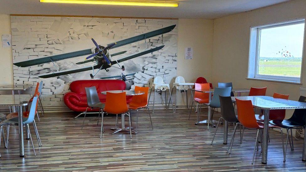 A wall mural depicting a biplane appearing to burst through a brick wall. The room has a brown laminate flood and several tables with metal chairs in it.