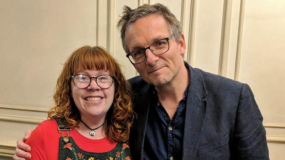 Claire Yates, a woman with red hair and glasses wearing a red top and dungarees, with Dr Michael Mosley