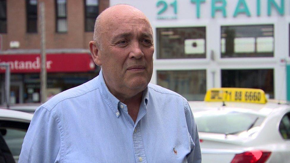 Kieran Kennedy is wearing a blue shirt and standing in the main street of Strabane. Shops and a bank can be seen in the background. He is standing beside a taxi.