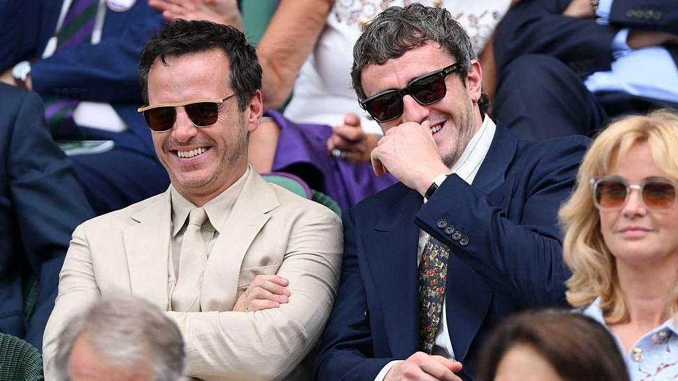 Andrew Scott and Paul Mescal sit side-by-side at the royal box at Wimbledon. Scott wears a beige coloured suit and is smiling, while Mescal wears a navy coloured suit and laughs as his hand slightly covers his face. Both men are wearing sunglasses.