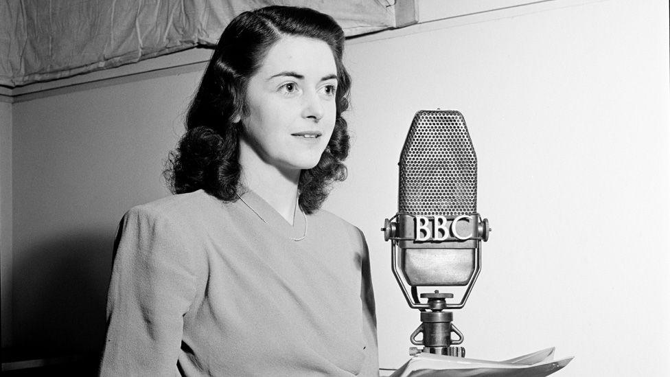 Black and white photo of Spencer standing in front of an old-fashioned BBC microphone holding a script