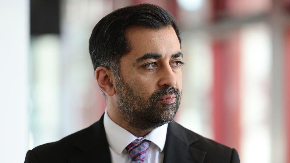 Humza Yousaf, with dark hair, a beard and wearing a black suit and purple tie, looks to his left in a close-up shot 