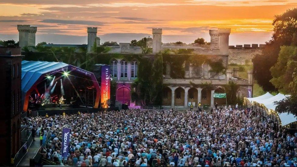 An outdoor concert is happening in front of a historic, ivy-covered building with towers and arched windows. A brightly lit stage is set up on the left, and a large crowd fills the open space, watching the performers. The sky glows with sunset colors of oranges and pinks.