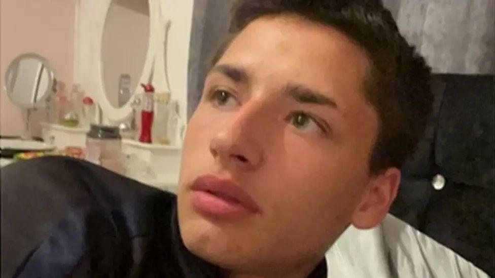 Cameron Bailey leaning back on what looks like a bed, he is looking up sideways. He has short dark hair and there is a mirror dressing table in the background.