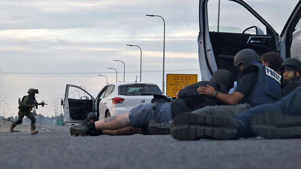 Journalists take cover behind cars as Israeli soldiers take position during clashes with Palestinian fighters near the Gevim Kibbutz, close to the border with Gaza on 7 October, 2023