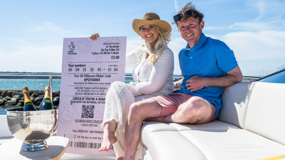 A man and a woman sat side-by-side on a boat holding a huge EuroMillions receipt 