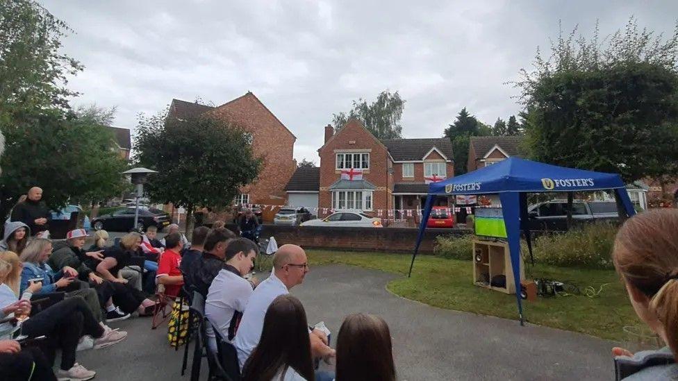 About a dozen people - some in football shirts - sit on garden chairs in two rows as they watch the match in the street. They are facing a television on a stand which is covered by a blue gazebo