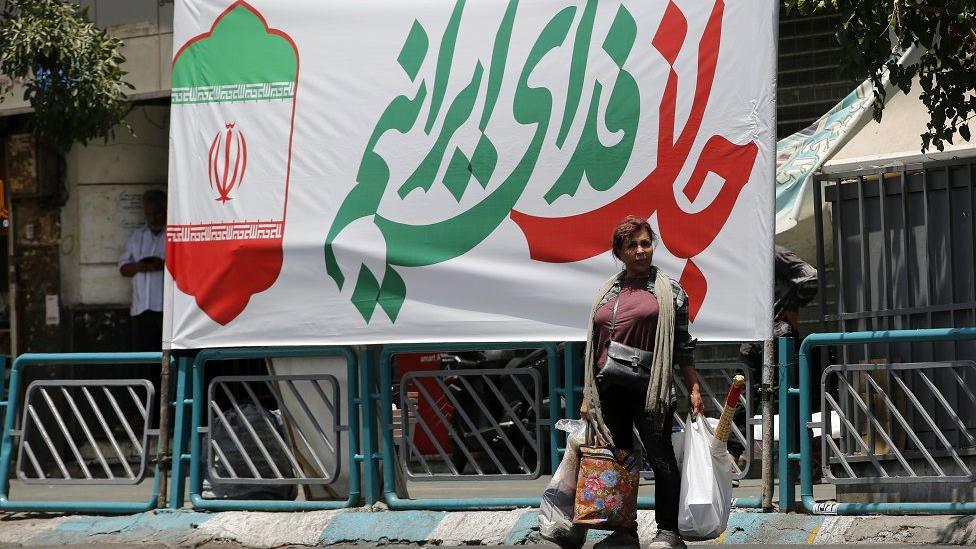 A woman holds bags in each hand as she stands at the side of the road.