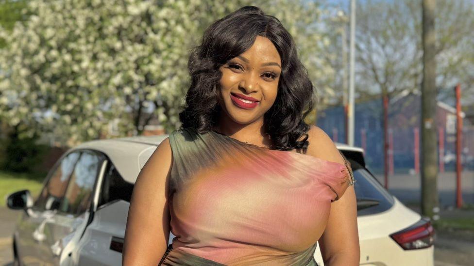 A woman with dark, curly hair in a pink dress is stood smiling outdoors in front of a white car and some trees