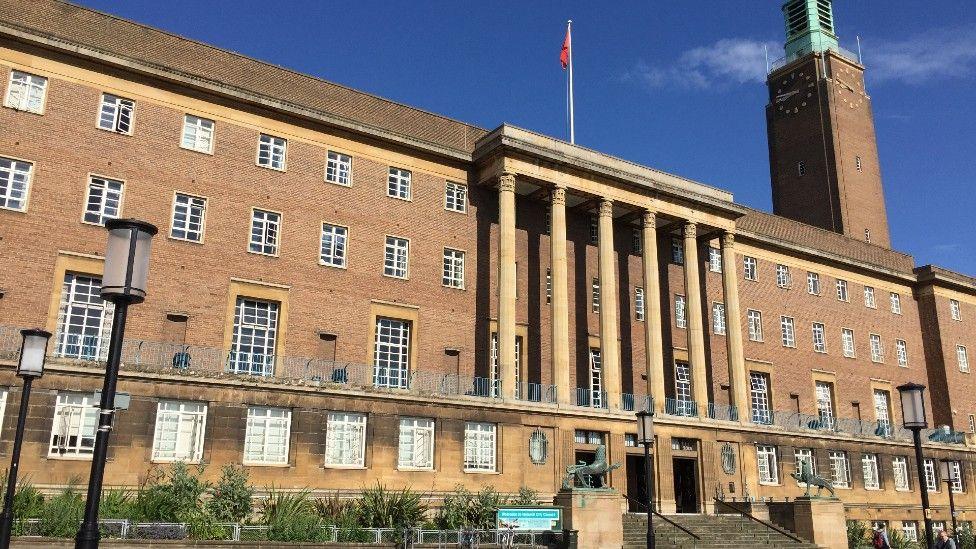 Norwich City Hall, a four-storey brick building with clock tower, columns at the front and a flag pole, set against a bright blue sky.