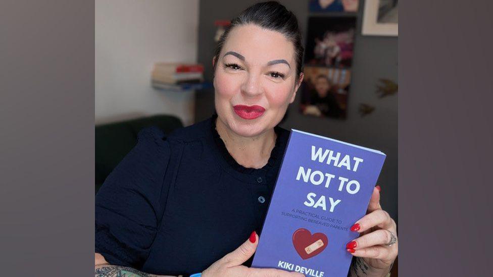 Kiki Deville smiles at the camera holding her book, which has a purple cover and is illustrated with a love-heart affixed with a sticking plaster