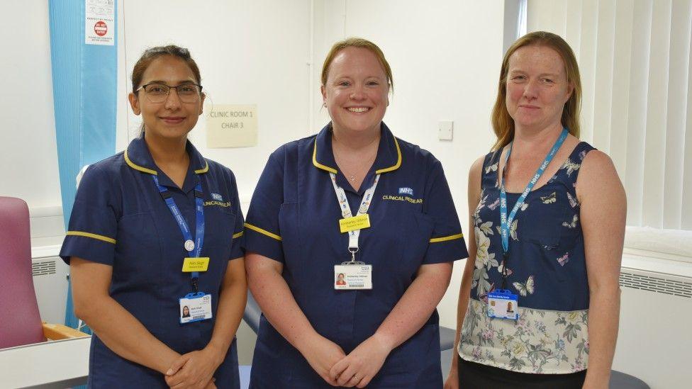 Three women wearing blue scrubs are standing next to each other, smiling at the camera. They are in a clinic room.