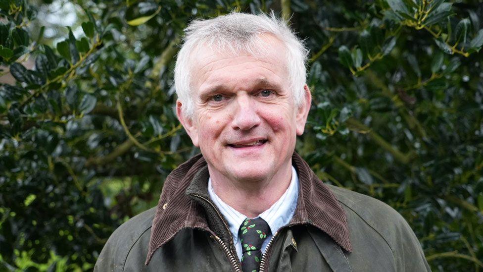 Nick Coller standing in front of a dense holly tree. He has short white hair and is wearing a green waxed jacket over a fleece and a white shirt with a black tie with pictures of holly on it