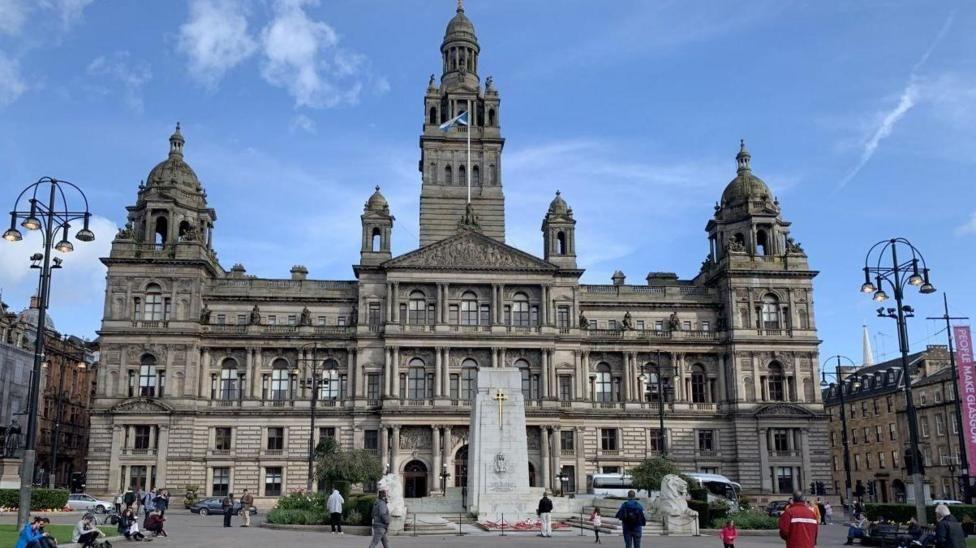A large ornate building with a public square in front of it. The sky is blue and there are people in the square.