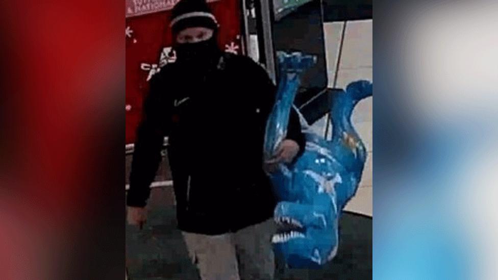 CCTV image of man in black scarf, and black hat, with only his eyes visible, and a black jacket and light grey trousers. In his left arm he is carrying a sculpture of a T-Rex, which is painted blue 