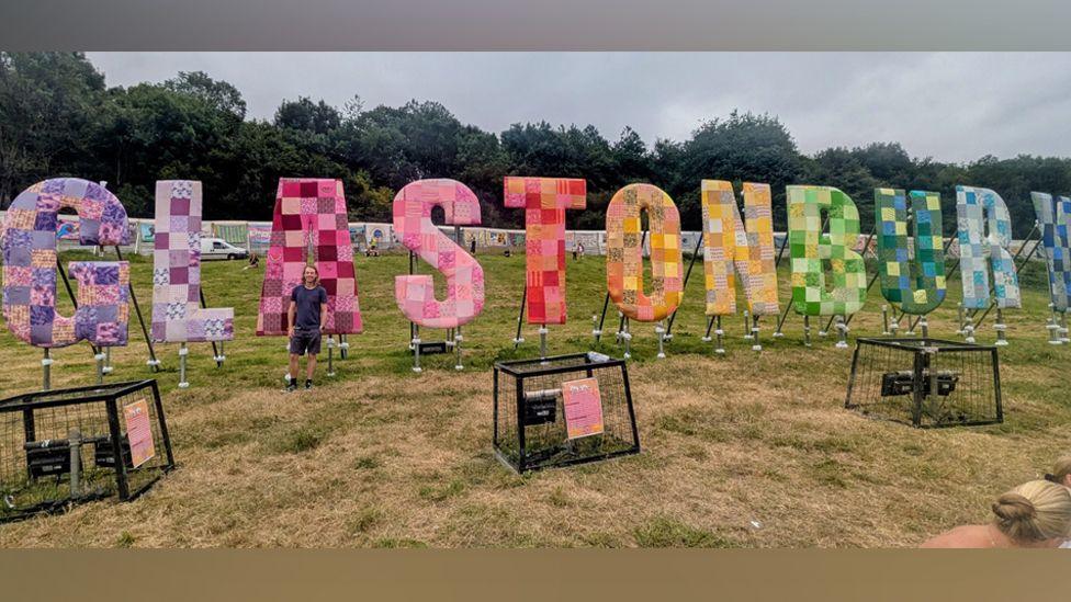 Lewis standing in front of a row of large letters that spell out Glastonbury, that is in lots of different colours. He is wearing shorts and a top. He has his hands in hit pockets.