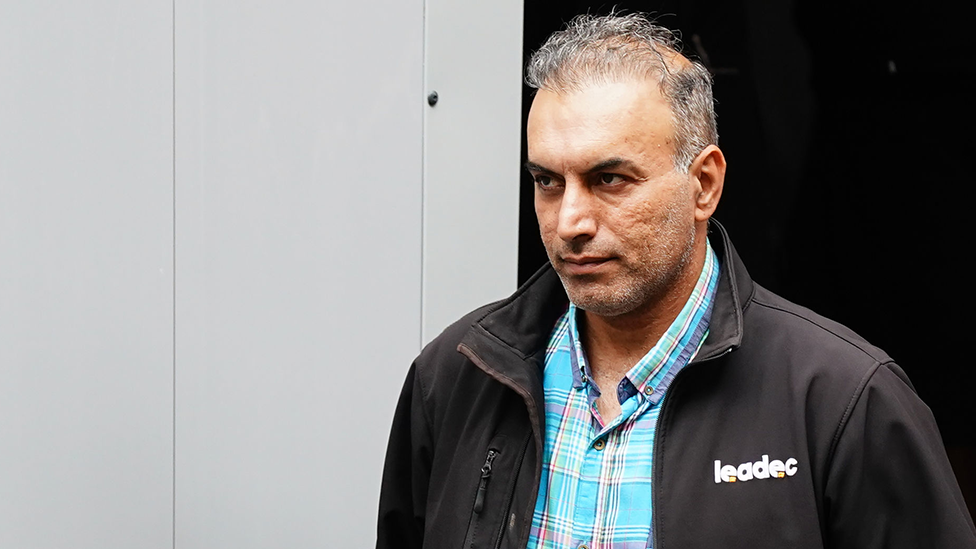 A man in a blue checked shirt and black jacket walking out of a court with a white and black background