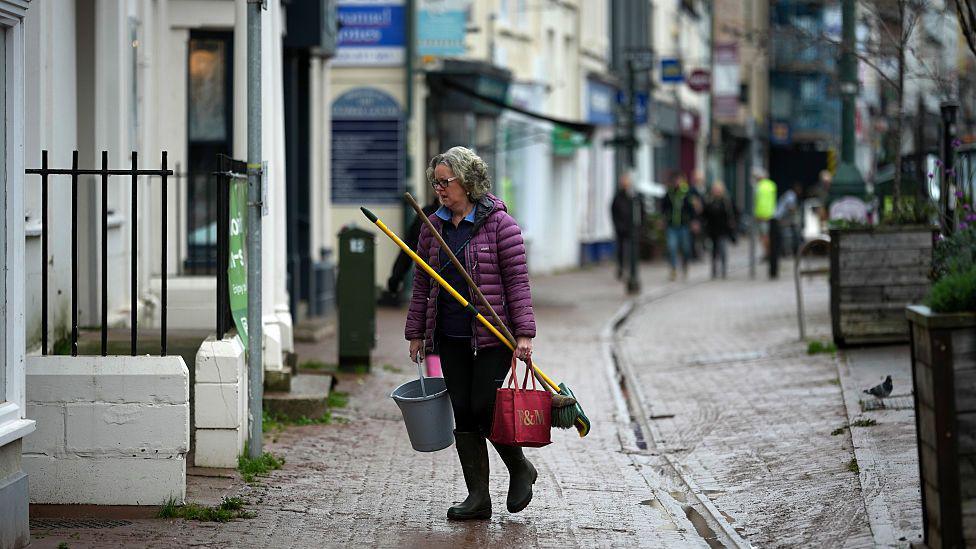 Menyw yn cerdded ar y stryd gan gario bwced a brws, yn ei hesgidiau glaw