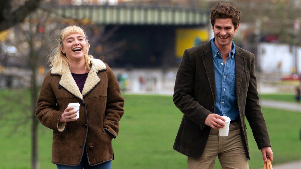 Florence Pugh and Andrew Garfield in We Live In Time, seen walking in a park carrying takeaway coffee cups