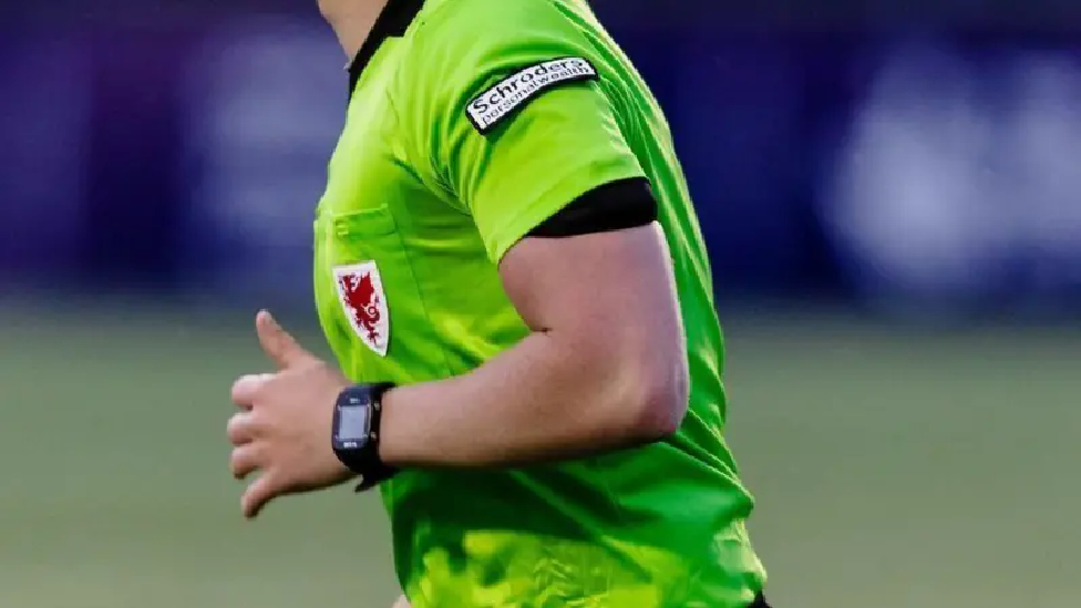 A man in a green referee shirt with a black watch and a Welsh emblem on the chest.