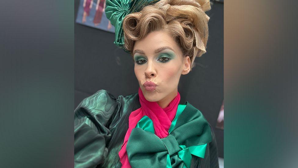 Lily Field wearing an emerald coloured costume, with an emerald bow and pink cravat.  She has blonde hair tied in a tall walnut whip style. She has emerald eye make up and is pursing her lips/blowing a kiss