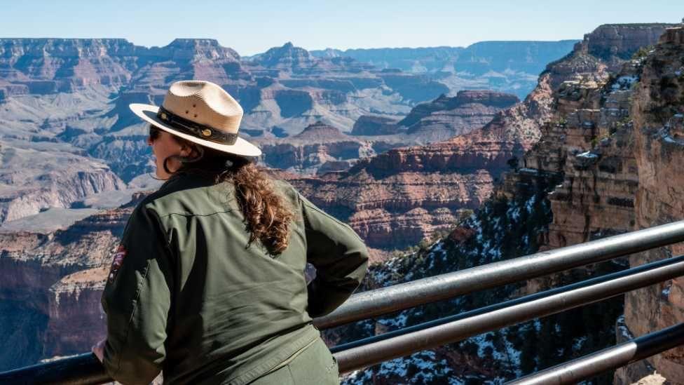 A national park ranger with a green jacket and a hat looks out into the snowy Grand Canyon National Park