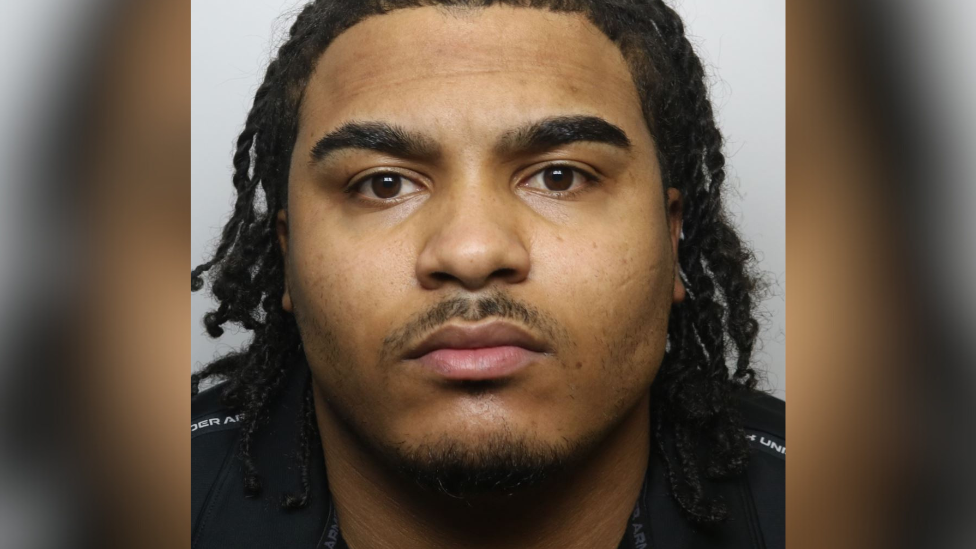 A police mugshot of Talbert Young, wearing a black t-shirt with his hair in shoulder-length braids.