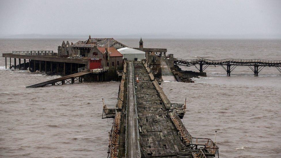 Birnbeck Pier which has fallen into disrepair. The wooden platform bridge is rotten and the planks are broken. The sea is brown and rough and the sky is dark grey.