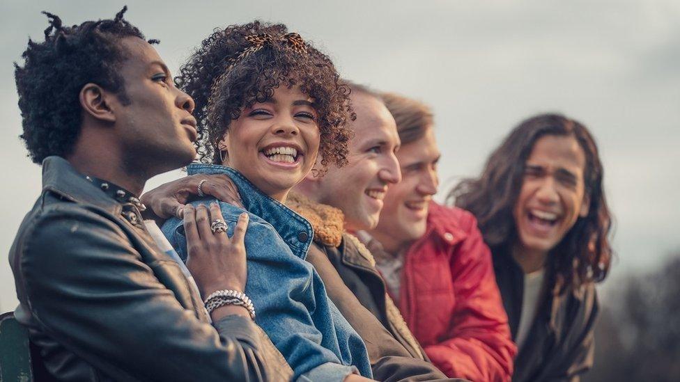 Five people sat in a row. They are all laughing, and have their arms round each other. Two of the men are wearing black jackets, one man is wearing a fluffy brown jacket and the fourth man wears a red jacket. The only woman is wearing a blue denim jacket and is the only only one looking at the camera.