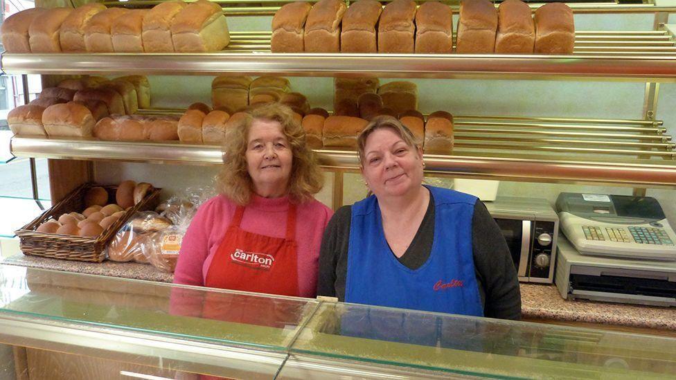 Bakery staff Brenda Jones and Karen Morgan standing in front of shelves of loaves and buns