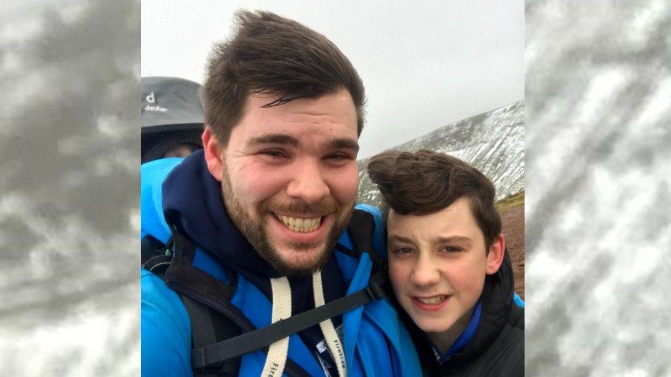 Michael Hall is wearing a bright blue coat and has his arm around his son Joshua who is wearing a black hoodie. The photo is a selfie and they are both smiling. 