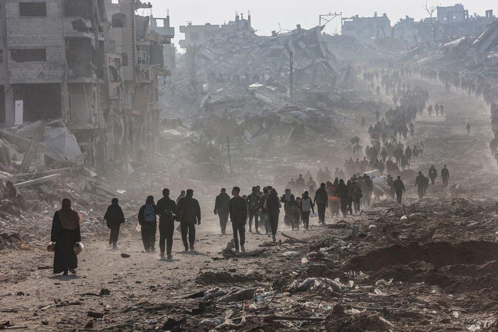 Scores of displaced Palestinians walk along a road in the Saftawi area of Jabalia, in the Gaza Strip, 19 January 2025.  A crowd of people in silhouette  walk into the distance, amongst the rubble of destroyed buildings.