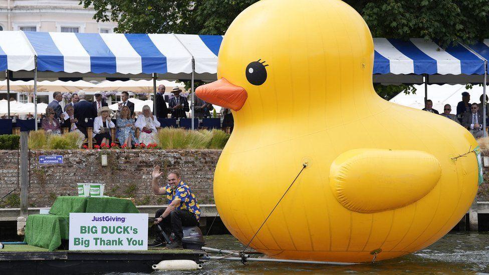 Giant inflatable yellow duck being towed on the Thames by a man on a small barge waving wearing a yellow duck printed blue shirt