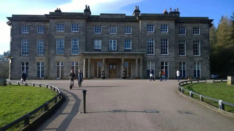 Haigh Hall, a stone manor house with several wings and levels seen from a large path where people are walking their dogs on a sunny day. 