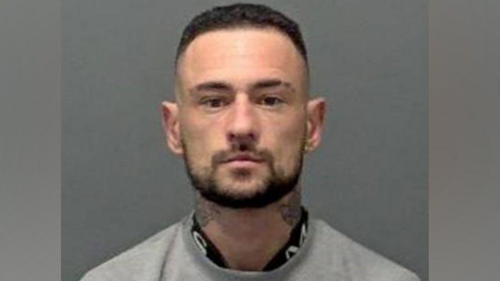 A police custody image of Kieran Humphreys: a man with a dark hair and a dark beard looking straight ahead. He has tattoos on his neck, is wearing a grey top.
