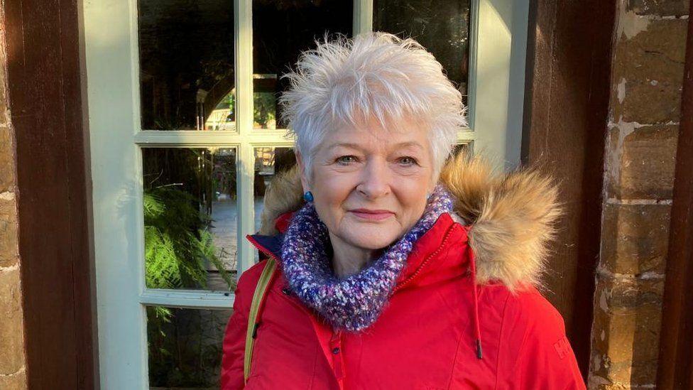 Wendy Hancock looking at the camera, with a door behind her. She has white hair and blue stud earrings in. She is wearing a red winter coat with a hood and a woollen scarf.