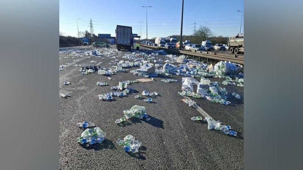 Multiple water bottles in clear and blue packaging are strewn across the motorway. A lorry and police car can be seen in the distance. Traffic travelling in the other direction appears to be queueing.