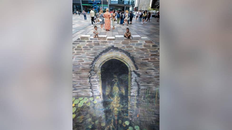 Artwork showing a water gate can be seen on the ground drawn in chalk. There is a water leading up to the gate with lily pads on the water in the art. Two children look at the art sitting on the ground.