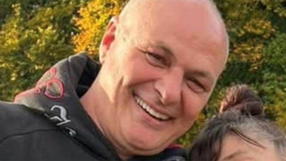 Close up of Wayne Putley smiling at the camera in front of some greenery. He has a shaved head and is wearing a dark hoodie