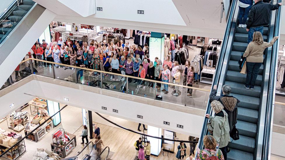 Choir singers perform on a John Lewis balcony, between the escalators.