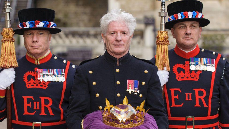 Bruno Peek in the gold buttoned uniform of the Queen's pageant master. He is flanked by two Yeoman warders at the Tower of London. He is holding a purple cushion with a crown surrounding the Jubilee Crystal Diamond. 