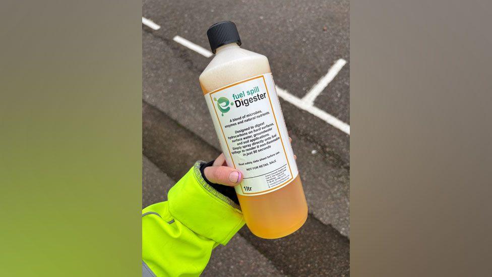 A hand holding a bottle of "fuel spill Digester" - an orange-coloured liqued in a white plastic bottle with a label on it.