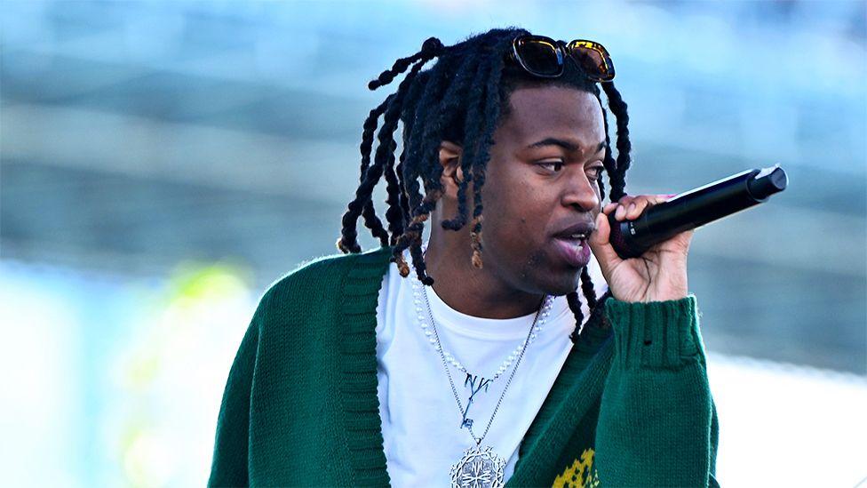 Jordan Adetunji performing in the US, wearing a green cardigan, while tshirt and silver chain. He is singing into a black microphone while looking to his left.