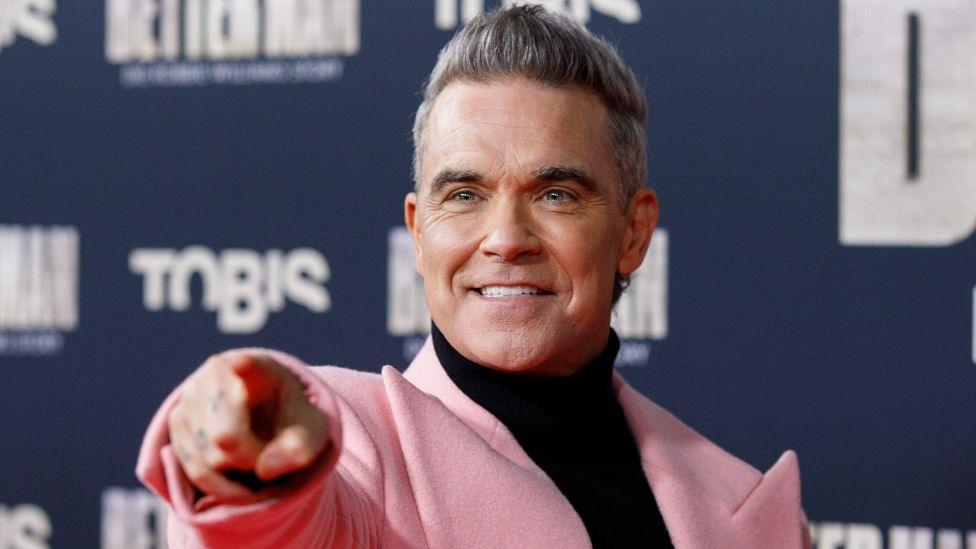 Robbie Williams attends the premiere of Better Man - The Robbie Williams Story at Cinedom on December 8, 2024, in Cologne, Germany. He is smiling on red carpet and pointing at the camera. He is wearing a pink jacket with huge lapels and a black polo-neck.